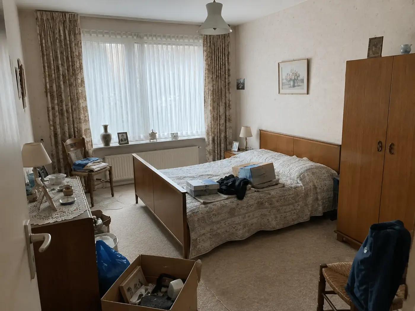 Slaapkamer vóór de ontruiming, met tweepersoonsbed, houten kledingkast, ladekast, stoelen en persoonlijke spullen.